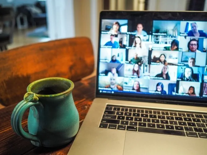 Laptop on a wooden desk showing a video call grid of students, beside a green ceramic mug
