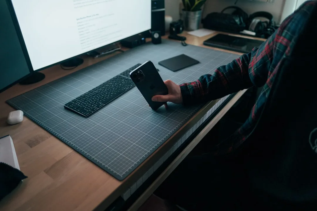 A person using a phone at a desk with a keyboard and monitor